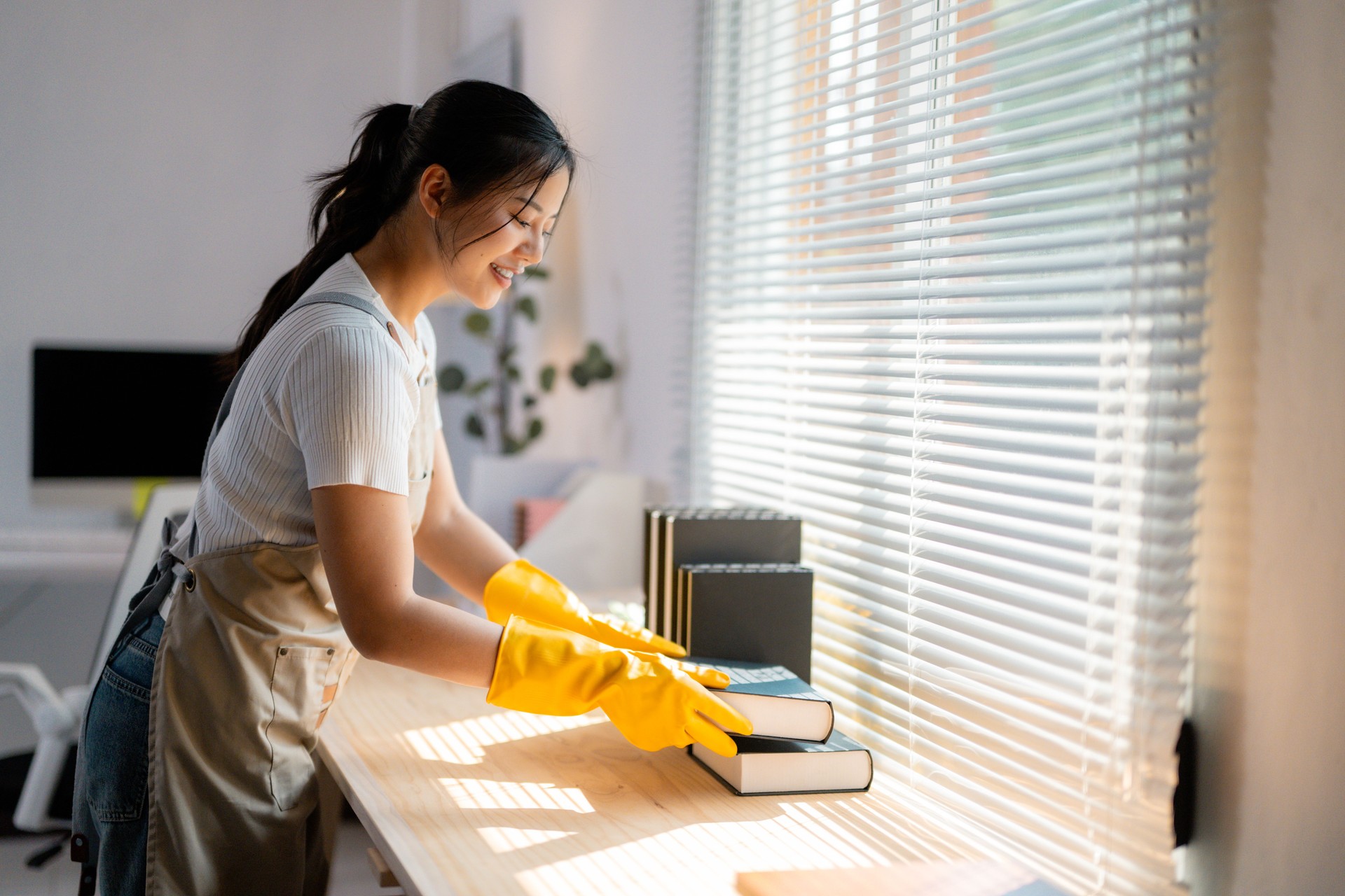 Smiling housekeeper wearing yellow protective gloves cleaning books on a wooden table in a sunlit room